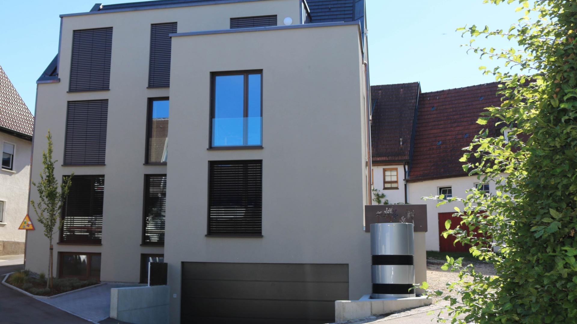 Ansicht eines modernen Mehrfamilienhauses in energieeffizienter Bauweise mit Wärmepumpe. Der geradlinig Baukörper wird durch große Fenster an der beigefarbenen Fassade strukturiert.